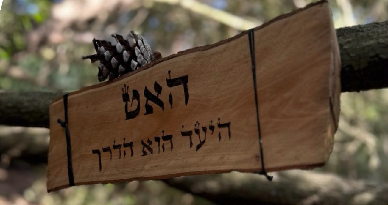 שלט עץ ועליו הכיתוב האט, היעד הוא הדרך, עם אצטרובל